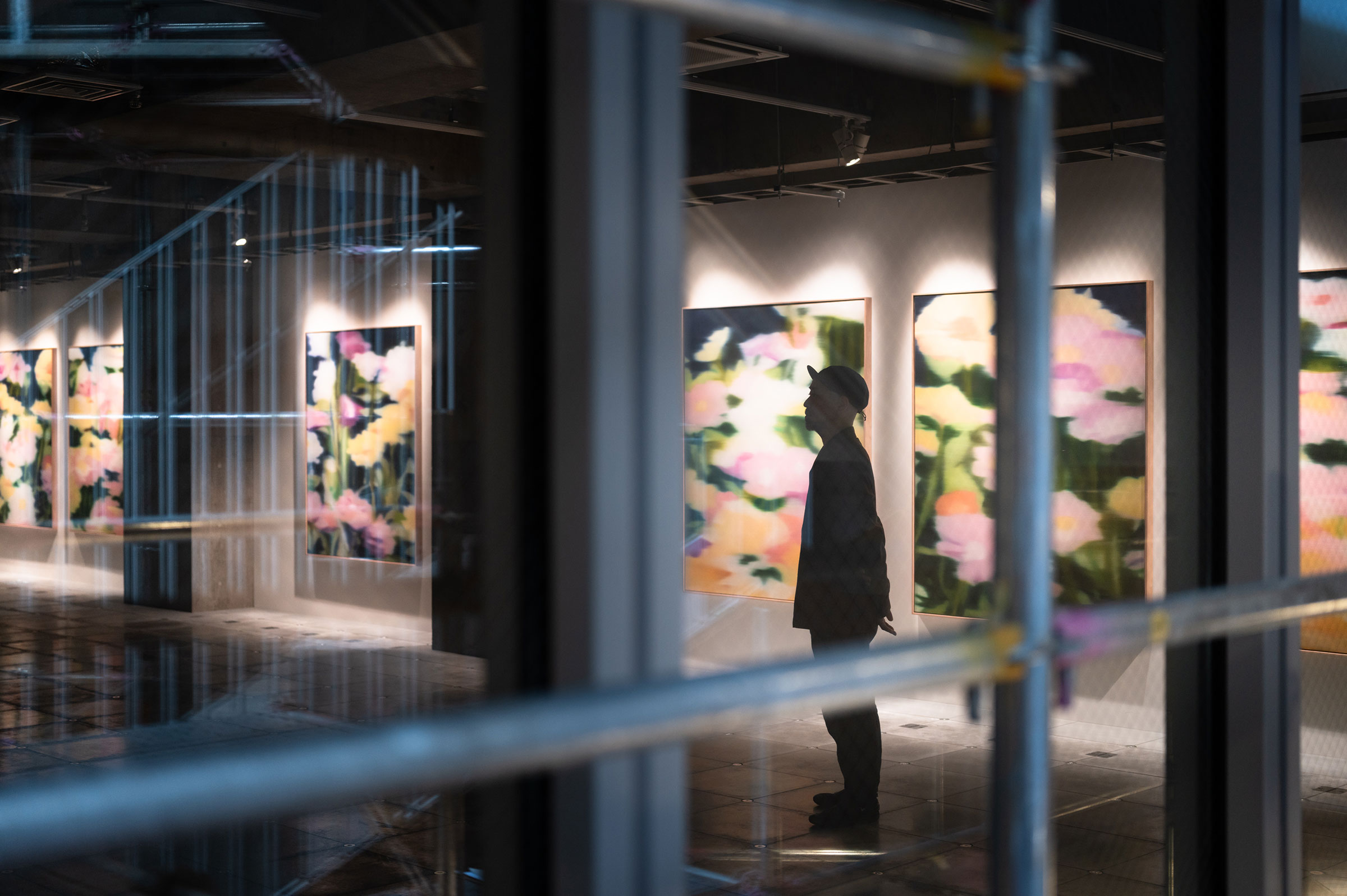 Exhibit. The silhouette of a person standing in front of softly colored artworks displayed on the wall.