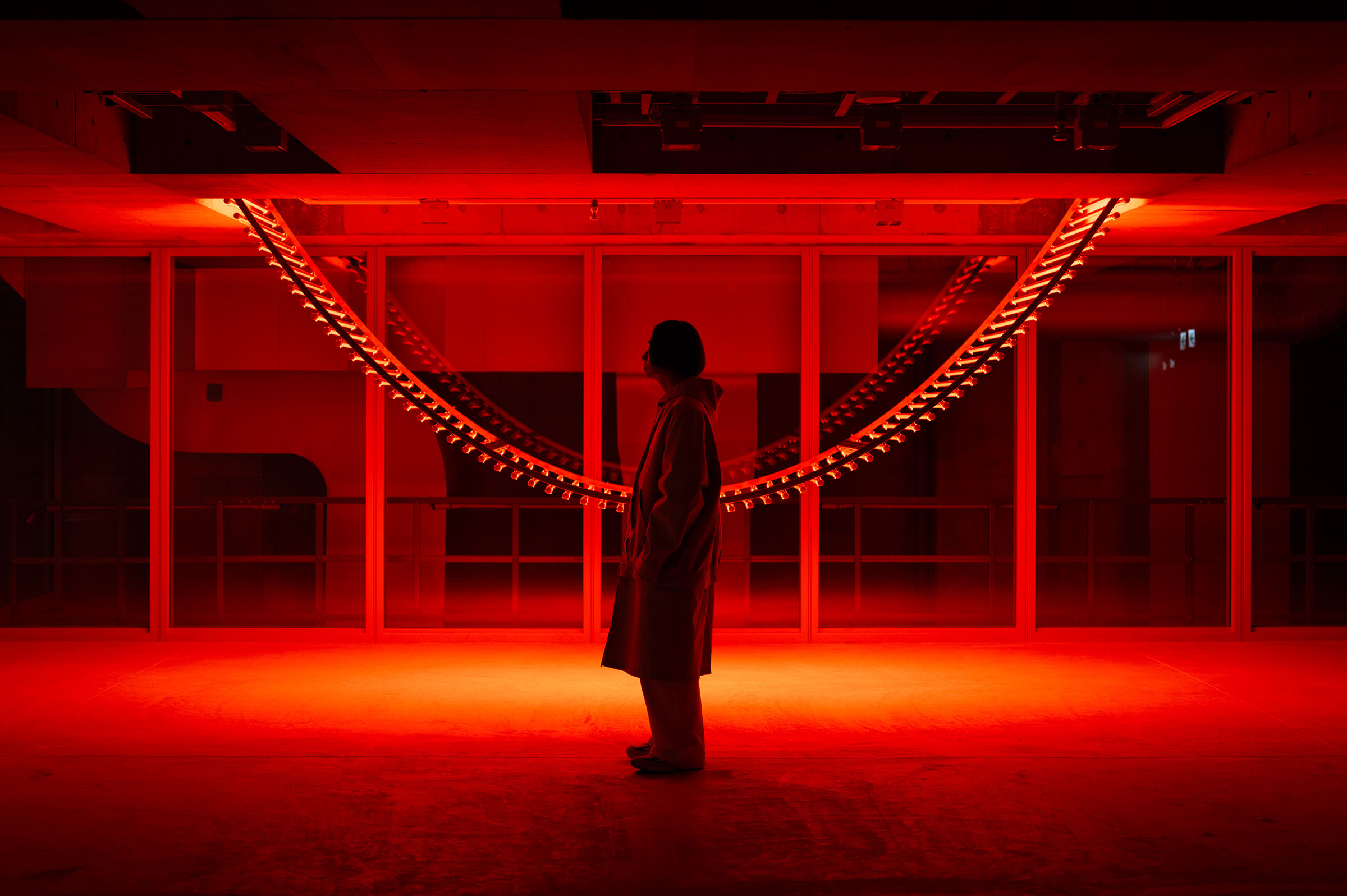  	Exhibit. The silhouette of a person standing in front of a piece illuminated by red lights.