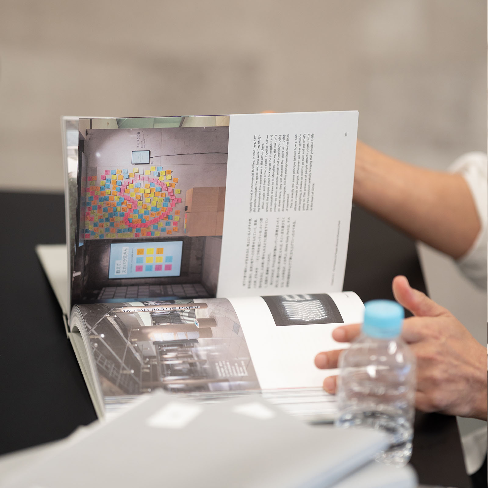 手前の人物がめくっている書籍『Document of Ginza Sony Park Project』の誌面アップ。左ページには「MUSIC IN THE PARK」と書かれた展示風景や色とりどりの付箋が貼られた壁の写真があり、右ページには英語と日本語のテキストと小さなモノクロ写真が掲載されている。