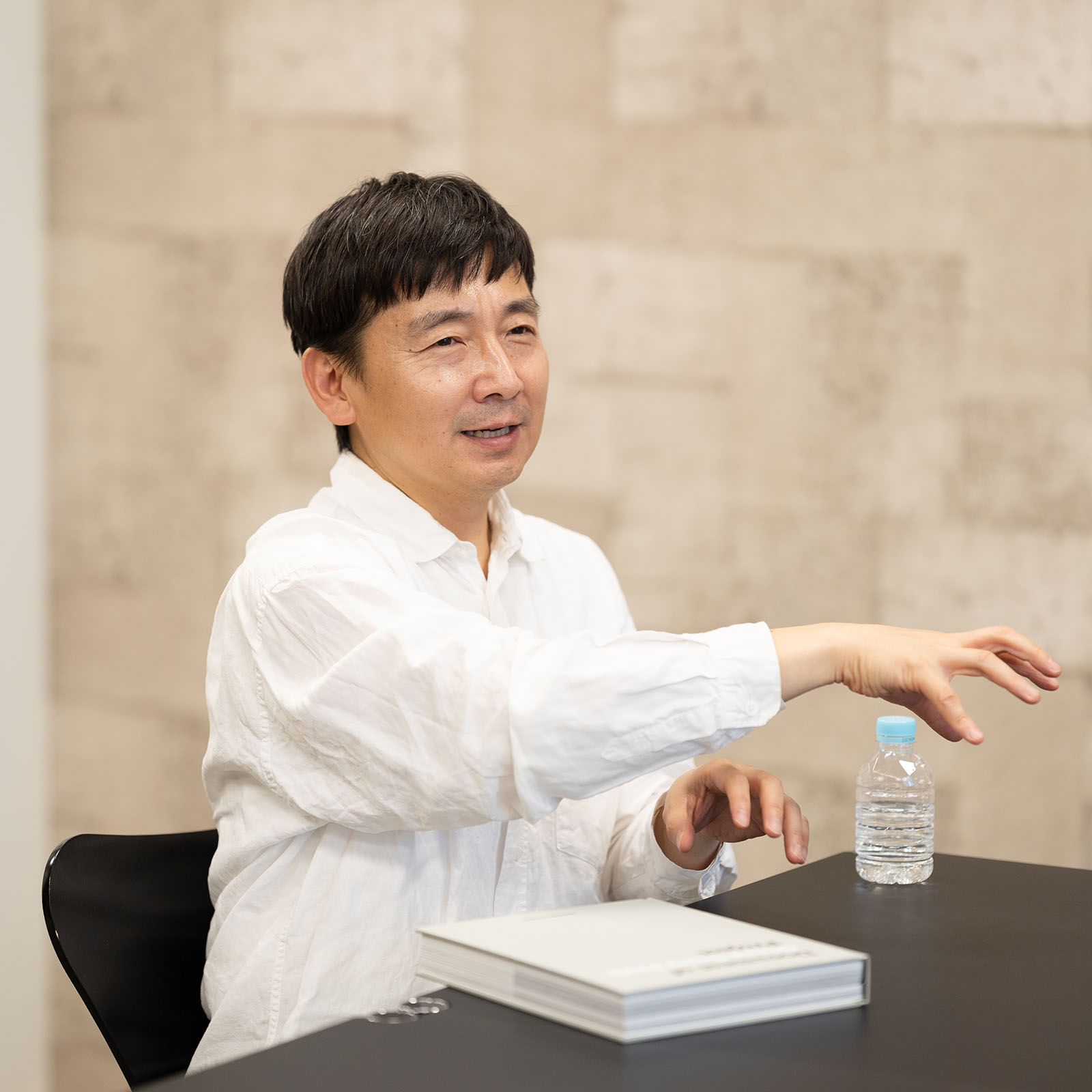 白いシャツを着た伊藤総研が、テーブルの上にある白い表紙の書籍『Document of Ginza Sony Park Project』の横で、右手を前に伸ばして身振り手振りを交えながら熱心に語っている様子。