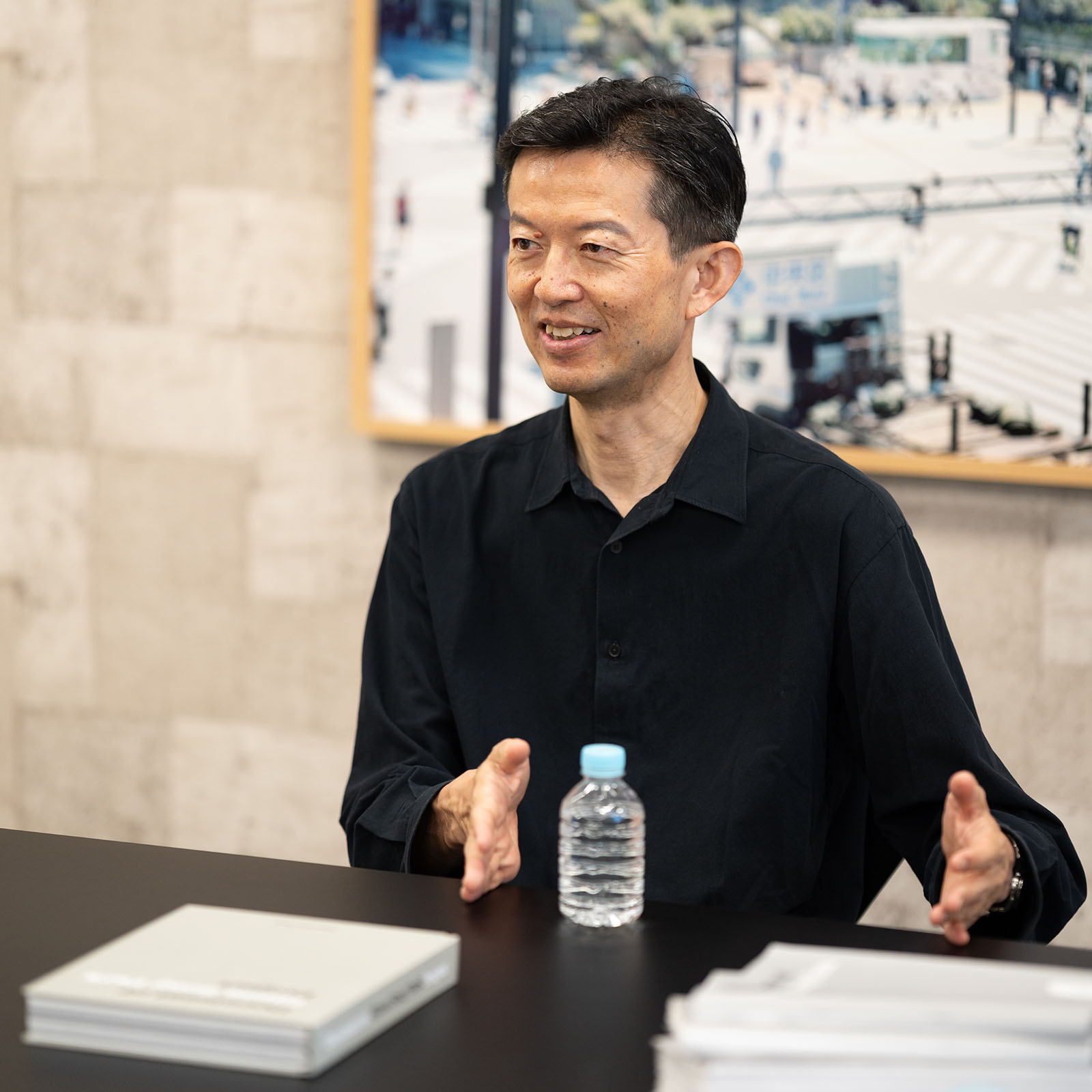 黒いシャツを着た永野大輔が、テーブルを挟んで笑顔で両手を広げながら鼎談相手に話しかけている。背景には銀座の街並みの写真が飾られており、テーブルには書籍と水ボトルが置かれている。