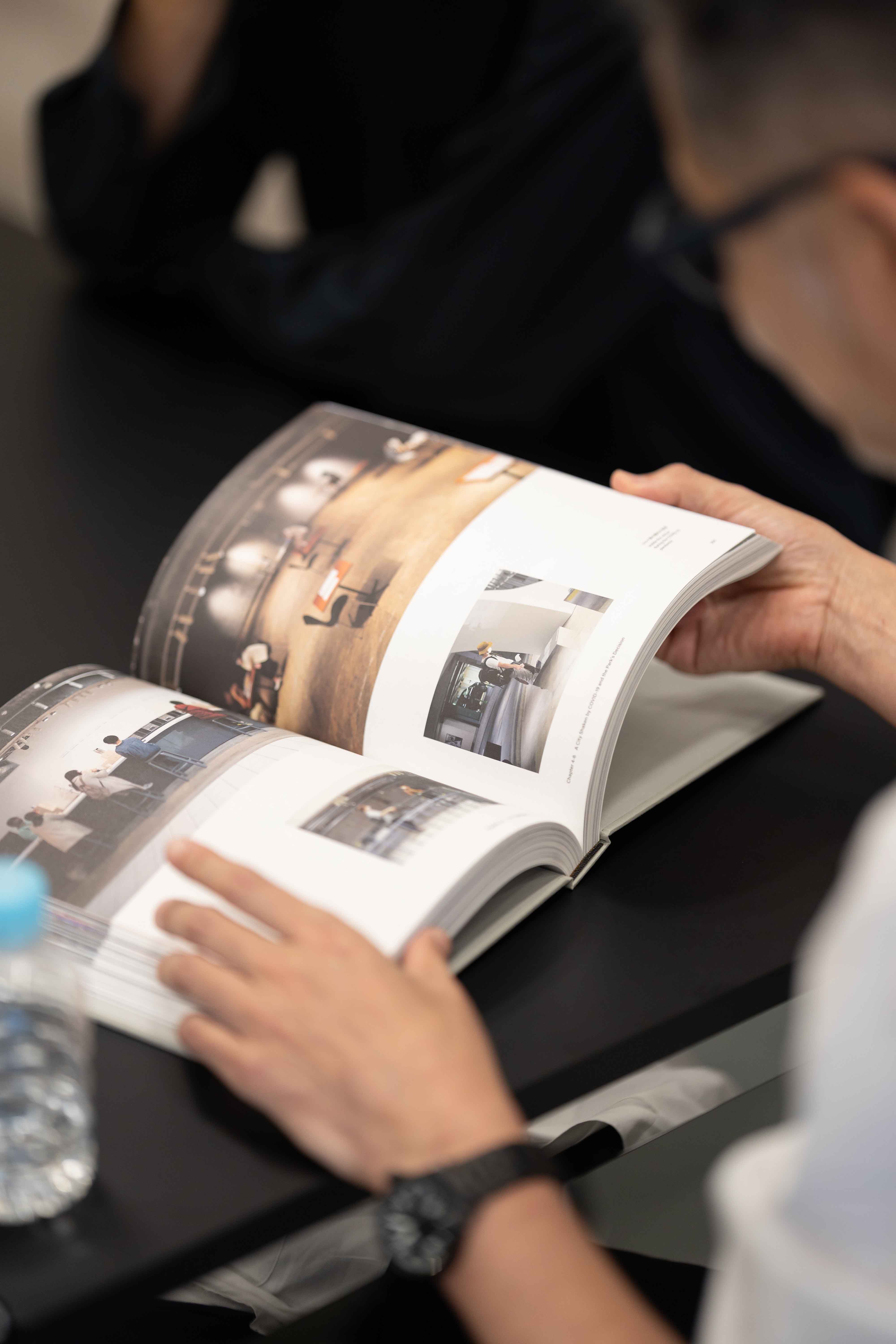テーブルの上で開かれた書籍『Document of Ginza Sony Park Project』のページを、手前の人物がめくっている手元のアップ。誌面には展示風景や空間の写真が多数掲載されている。