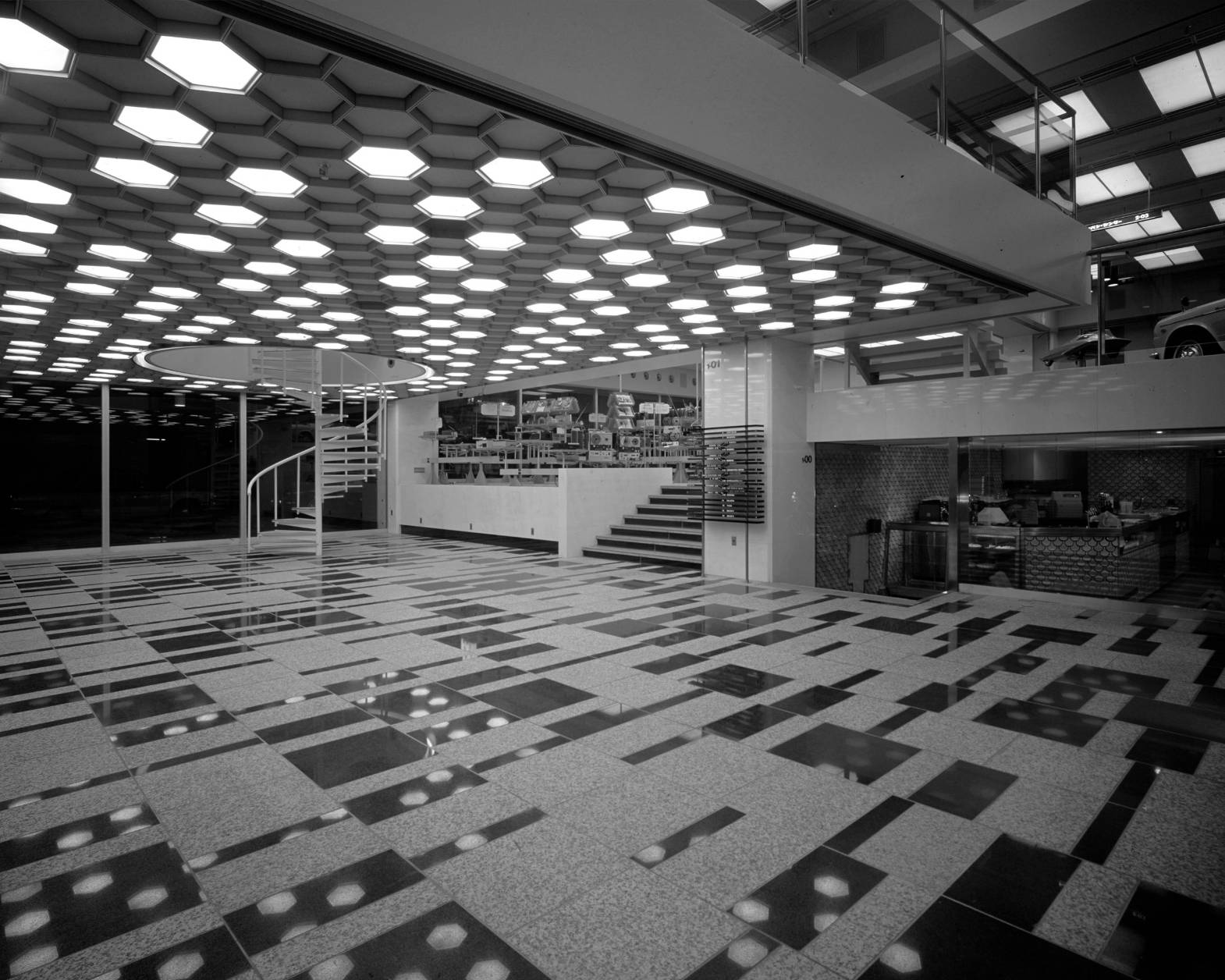 内観（モノトーン）。開業当時の1階エントランスホール。左奥には螺旋階段があり、天井にはハニカム構造の照明が広がっている。