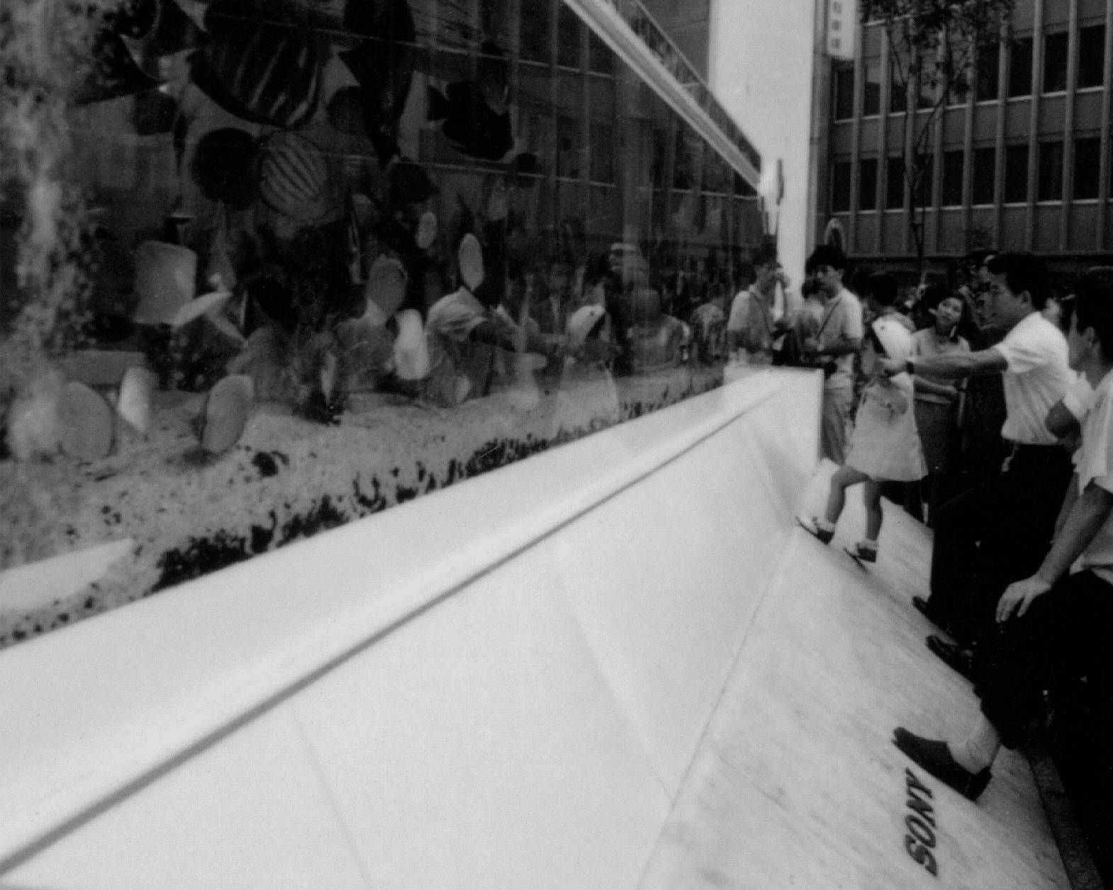 外観（モノトーン）。屋外に設置された巨大水槽に来街者が集まっている。小さな子どもを抱えて見せる父親の姿もある。