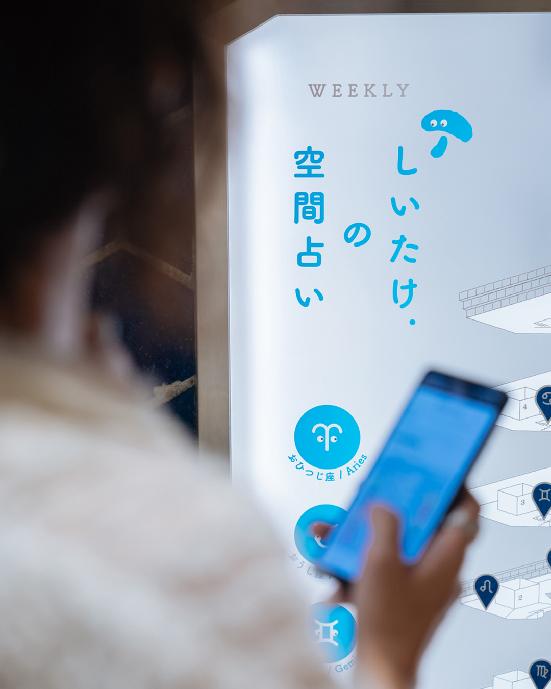 「しいたけ.の空間占い」のマップをスマートフォンで確認する来園者。ショーケースのマップには12星座のアイコンが並ぶ。