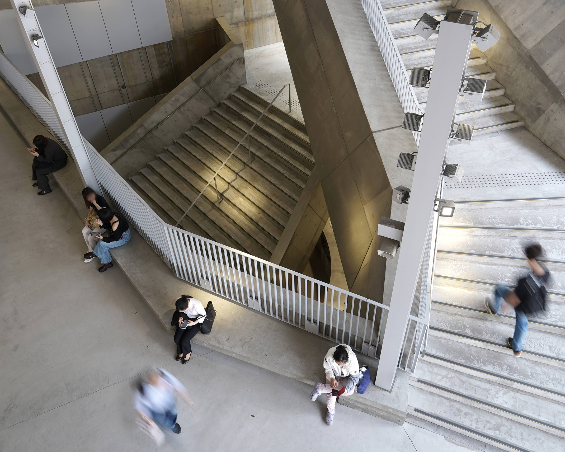 吹き抜けの階段空間を上から見下ろす。ベンチに座る人々と、階段を上り下りする人々の様子。