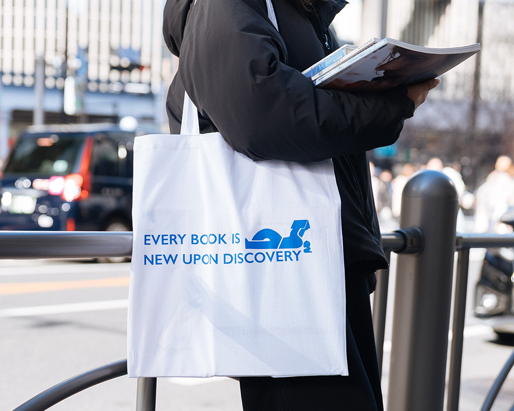 A visitor carries a tote bag purchased at Ginza Sony Park, holding a book in the city. The phrase “EVERY BOOK IS NEW UPON DISCOVERY” stands out clearly on the bag.