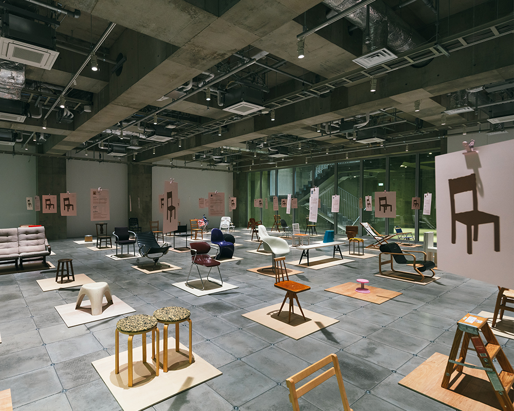 On the exhibition floor of Ginza Sony Park, diverse chairs stand on platforms beneath suspended panels, forming a dialogue within the concrete space.