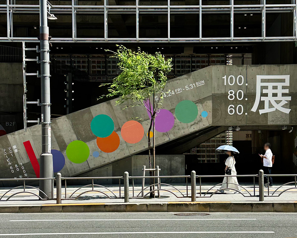 銀座の街路から見たGinza Sony Park。地下へ続くコンクリートのスロープ壁面に「100.80.60.展」の大型グラフィックが掲出され、歩道を行き交う人々の姿が見える。