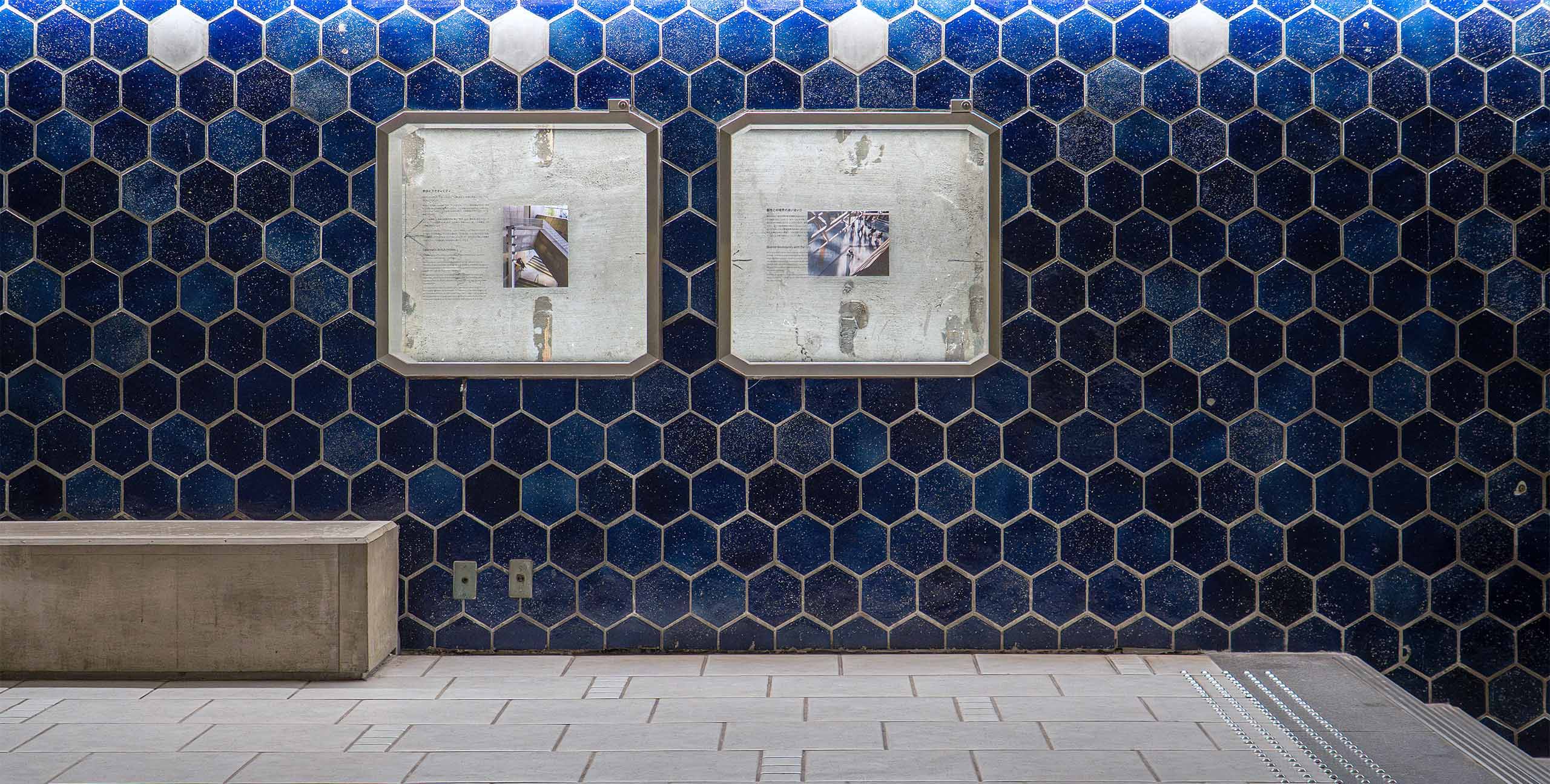 Two display panels are mounted on a wall of deep blue hexagonal tiles at Ginza Sony Park, with a concrete bench and tiled floor creating the quiet atmosphere of an underground passage.
