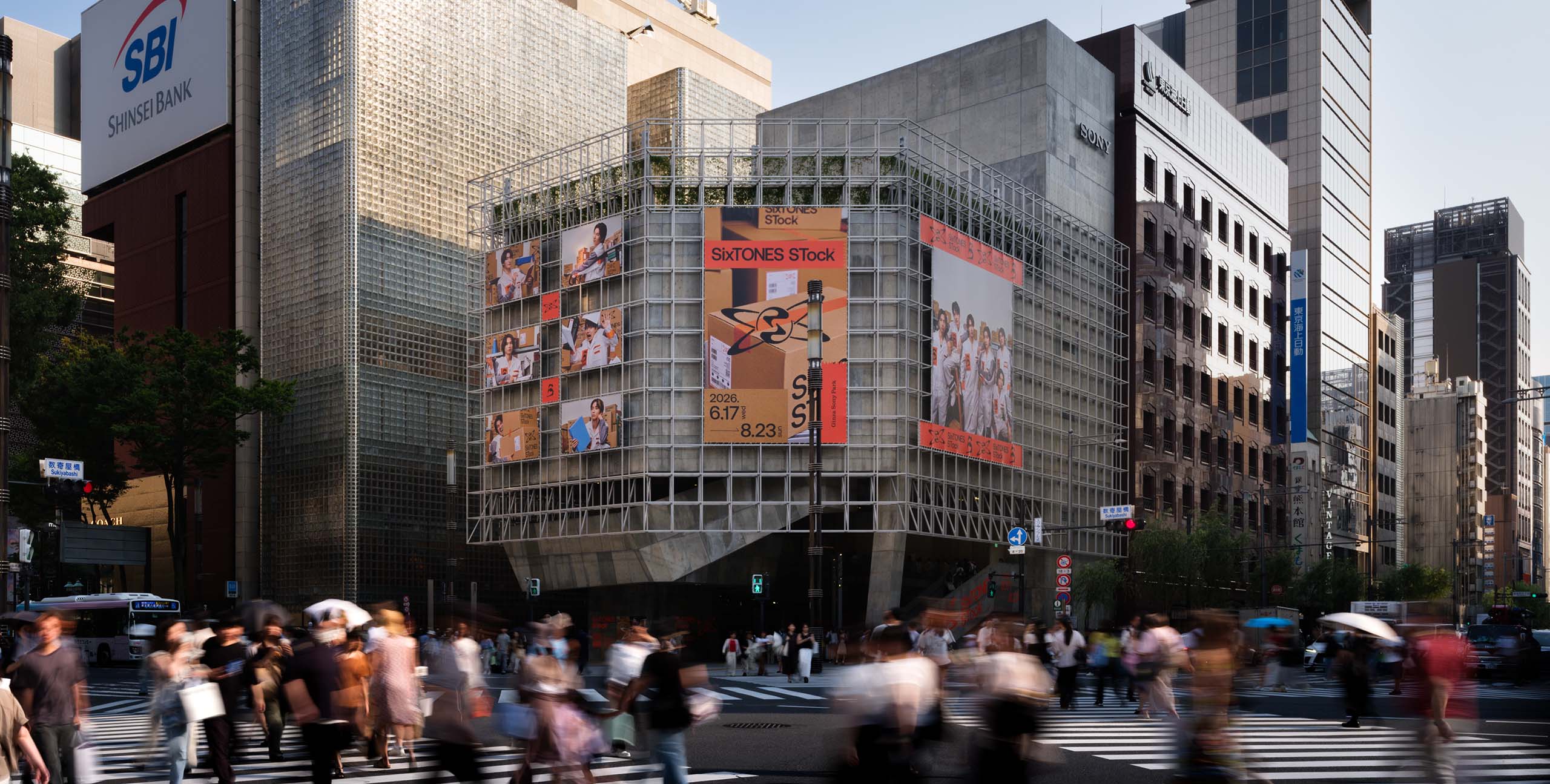 晴れた日のGinza Sony Parkの外観。金属フレームで覆われた建物の壁面には「SixTONES STock」の大型看板やメンバーの様々な写真、開催期間「2026.6.17 - 8.23」の文字が掲示されている。手前の横断歩道には、多くの人々が交差点を交錯して行き交う様子が動感をもって写っている。