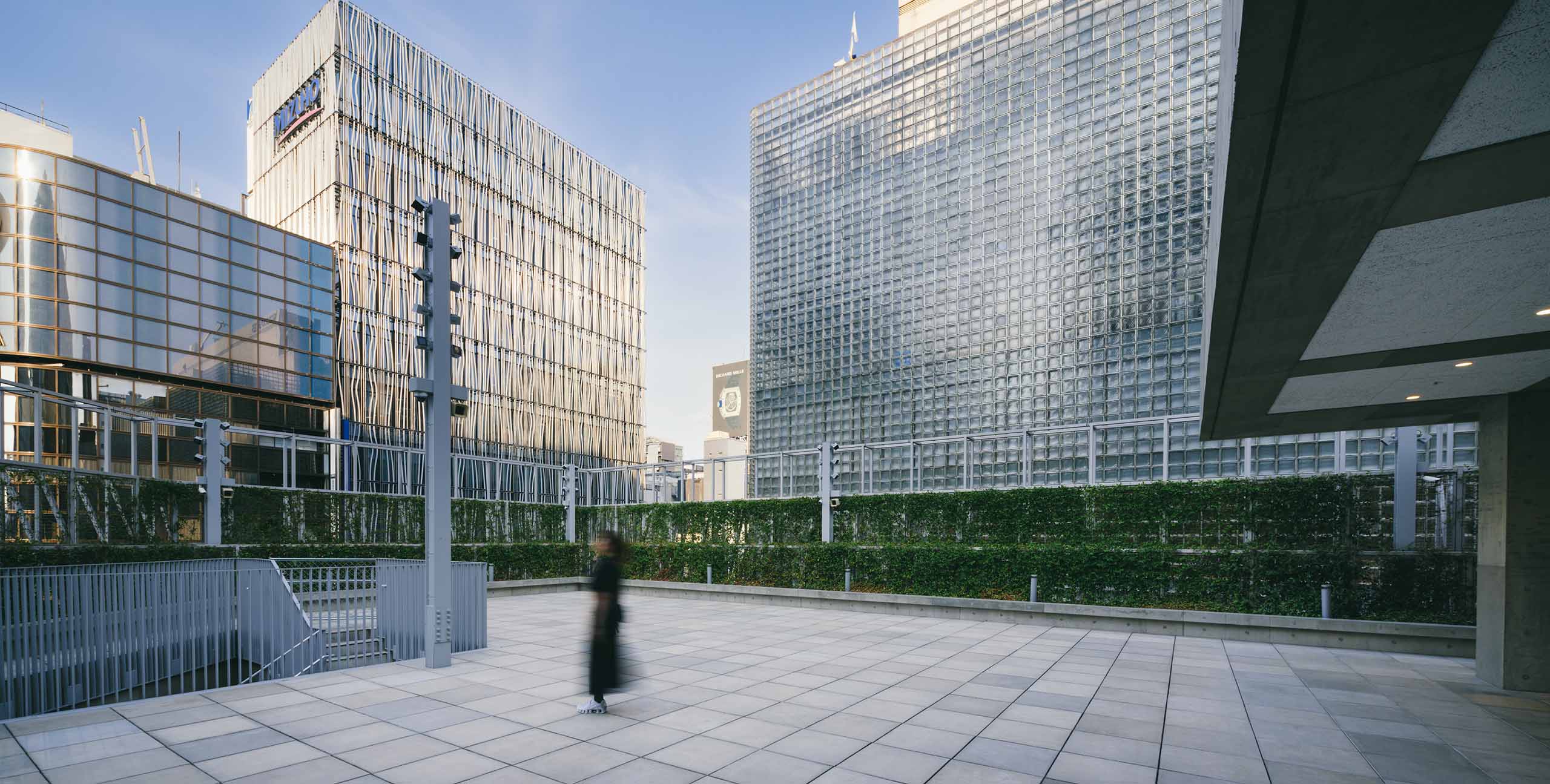 The rooftop terrace of Ginza Sony Park, featuring paved tiles, a green hedge, and a person walking in black clothes, framed by modern Ginza buildings in the background.