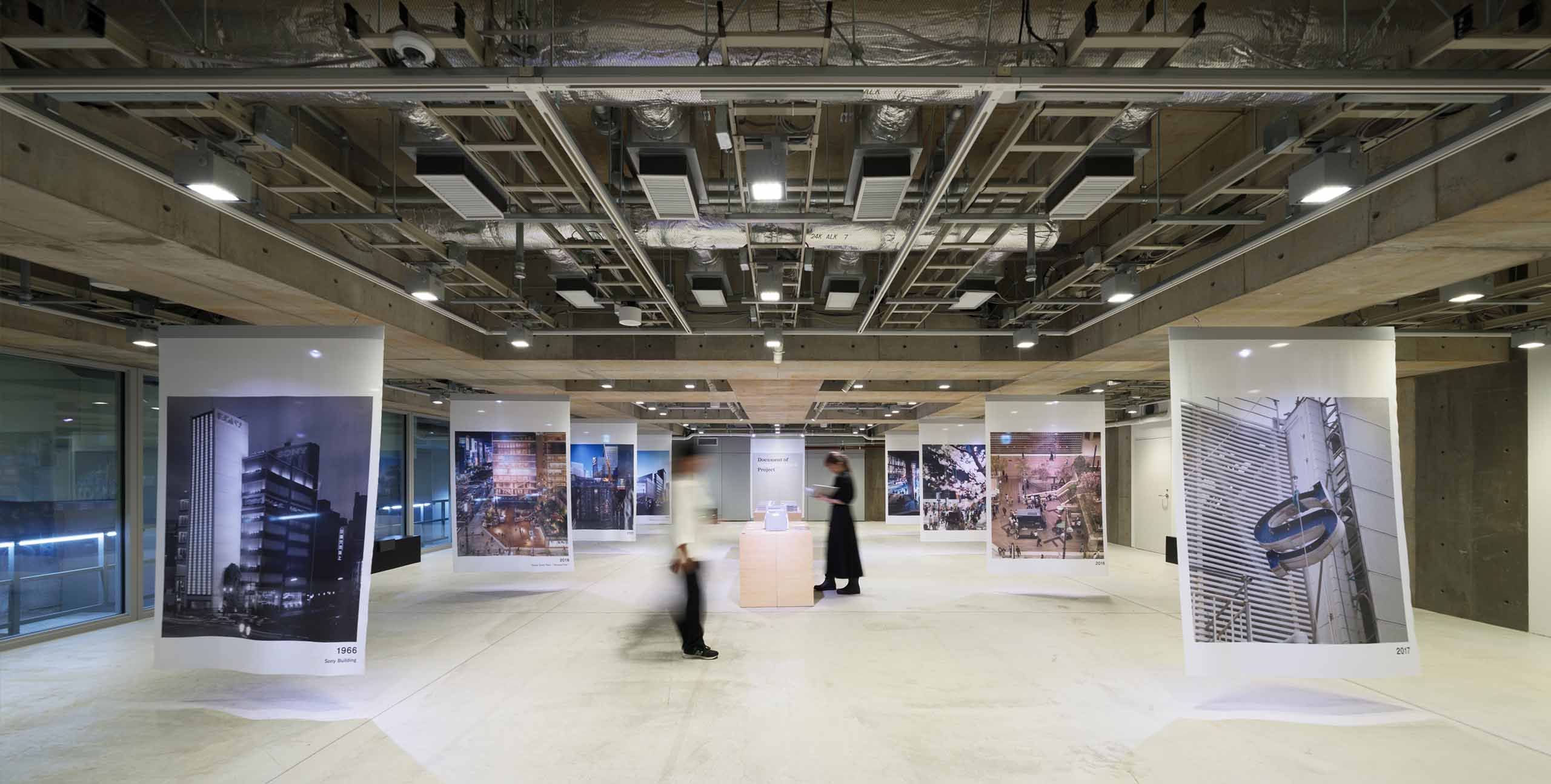 Ginza Sony Park B2で開催中の『Document of Ginza Sony Park Project』出版記念展示。コンクリートの空間にソニービル時代からParkの建設風景、日常の姿までを伝える巨大な写真幕が多数吊り下げられ、中央の展示台では来場者が書籍を手にとっている。