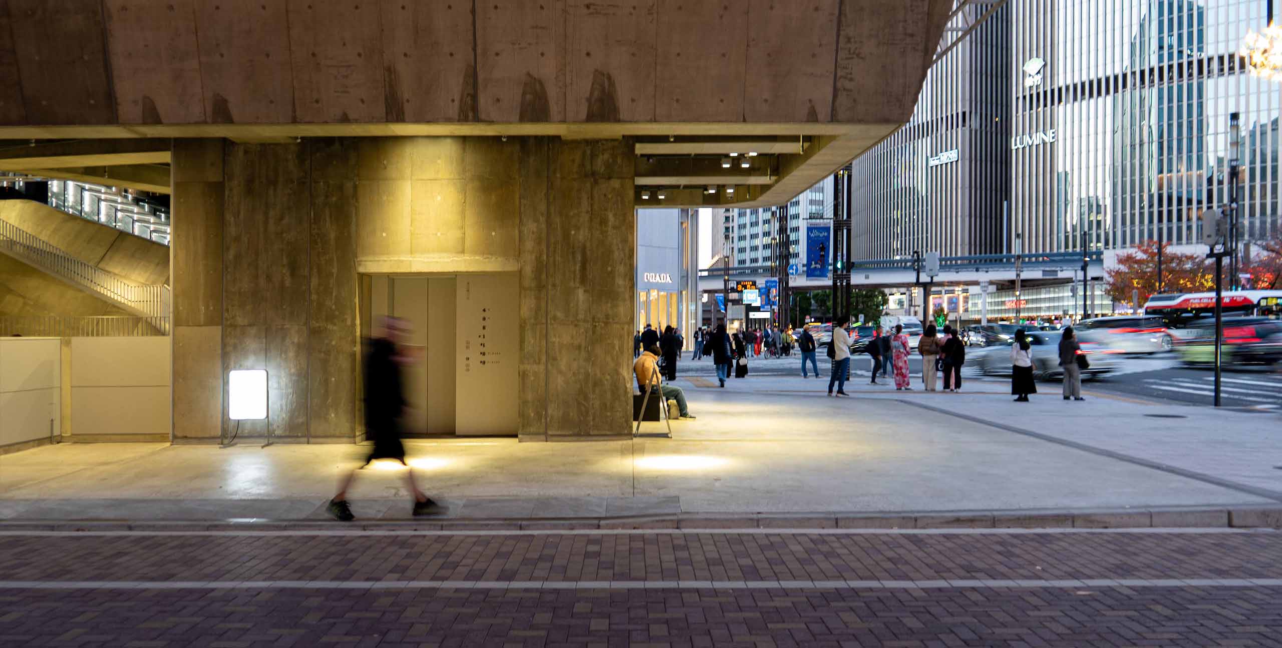 A night view of the ground floor of Ginza Sony Park. An open concrete structure protrudes onto the intersection, with a person walking and blurring in the foreground, an elevator and seated visitors behind them, and the traffic of Harumi-dori and the Ginza city skyline in the far background.